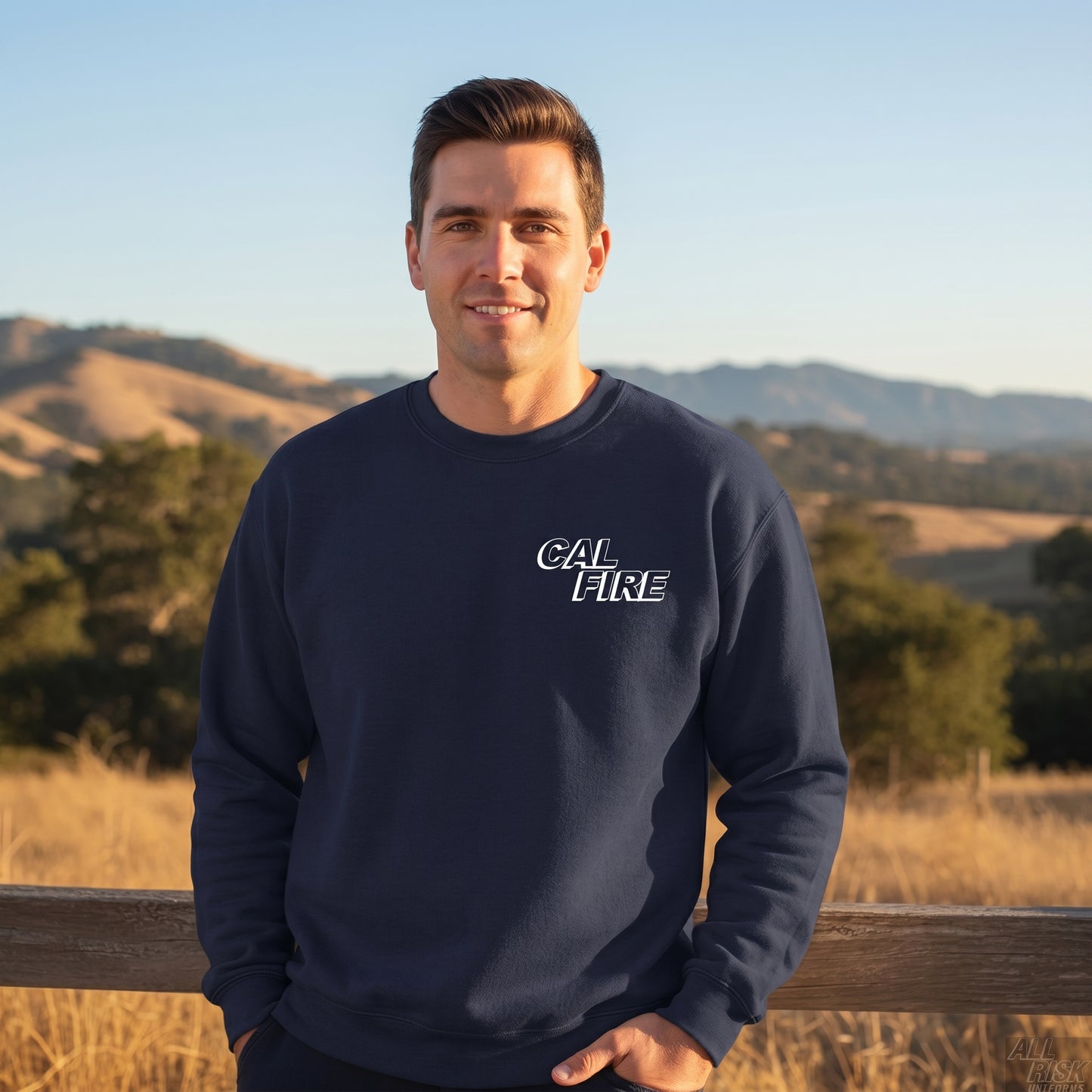 Lifestyle photo of a man wearing the navy CAL FIRE crewneck sweatshirt — leaning on a wooden ranch fence rail in golden California hills, oak trees and rolling grass-covered hills in the background. Off-duty / casual setting.