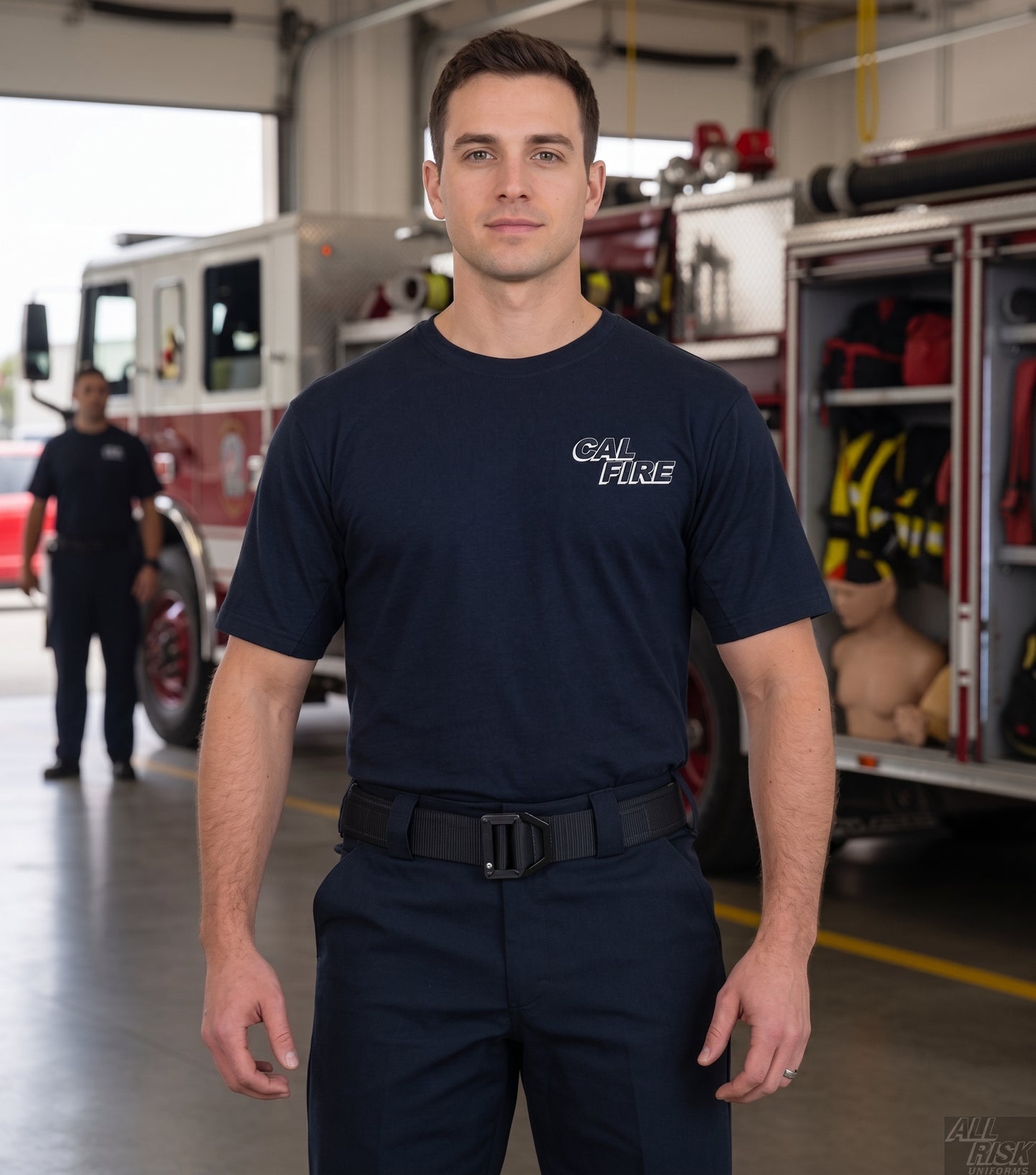CAL FIRE firefighter standing in a fire-station apparatus bay in front of a fire engine, wearing the navy First Tactical CAL FIRE cotton t-shirt with the white CAL FIRE italic wordmark on the left chest, navy duty trousers and belt; a second firefighter blurred in the background.