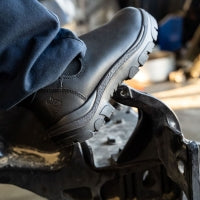 Crew member's feet in the black Steel Blue Hobart steel-toe slip-on chelsea boot — close-up of the side detail with elastic gusset and lugged outsole on a workshop floor.