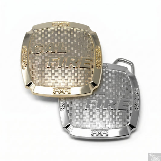 CAL FIRE belt buckle — gold-brass and silver versions side by side, each rectangular cushion shape with raised CAL FIRE serif lettering on a basket-weave-textured center field, decorative scrollwork at the side panels, and small ornamental frames at top and bottom centers.