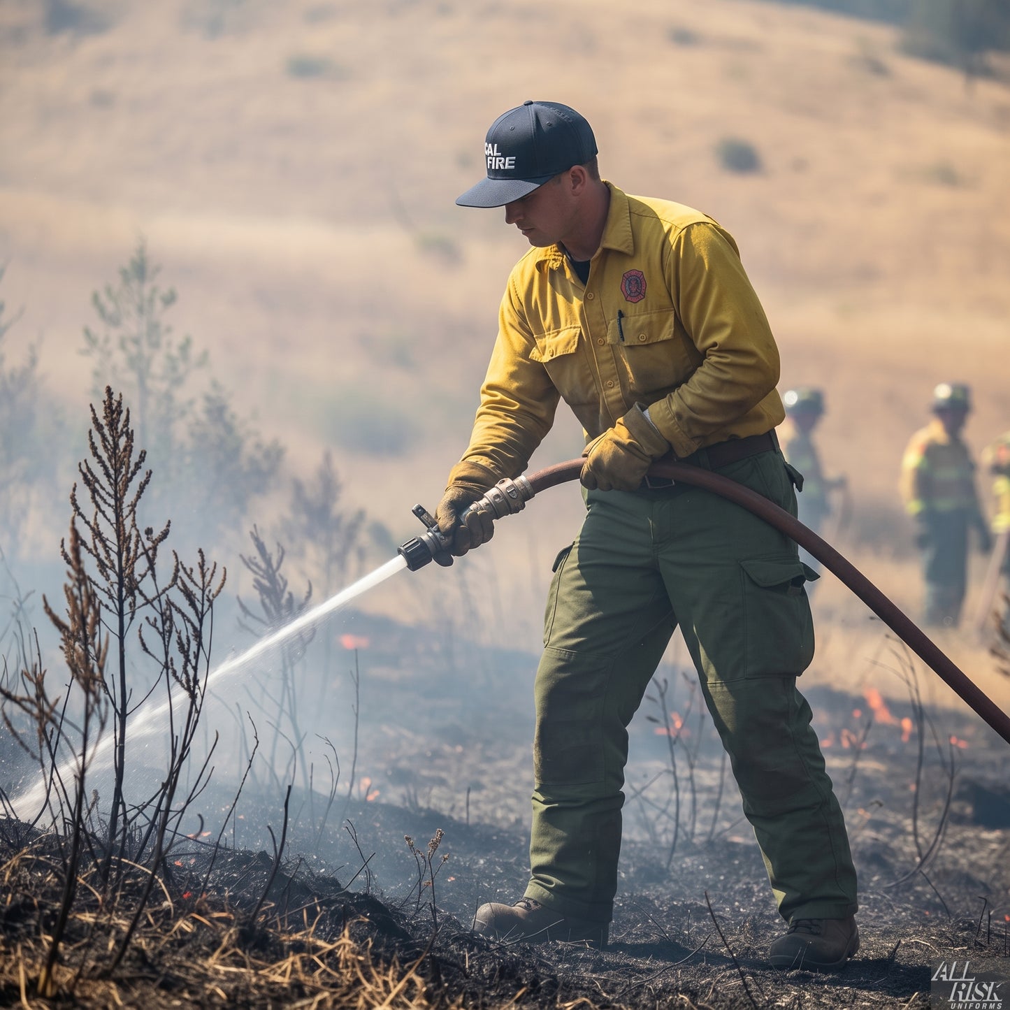 CAL FIRE wildland firefighter in a yellow Nomex brush shirt and green pants spraying a hose line on a grass fire while wearing the navy CAL FIRE Bucket Brigade cap, crew working behind.