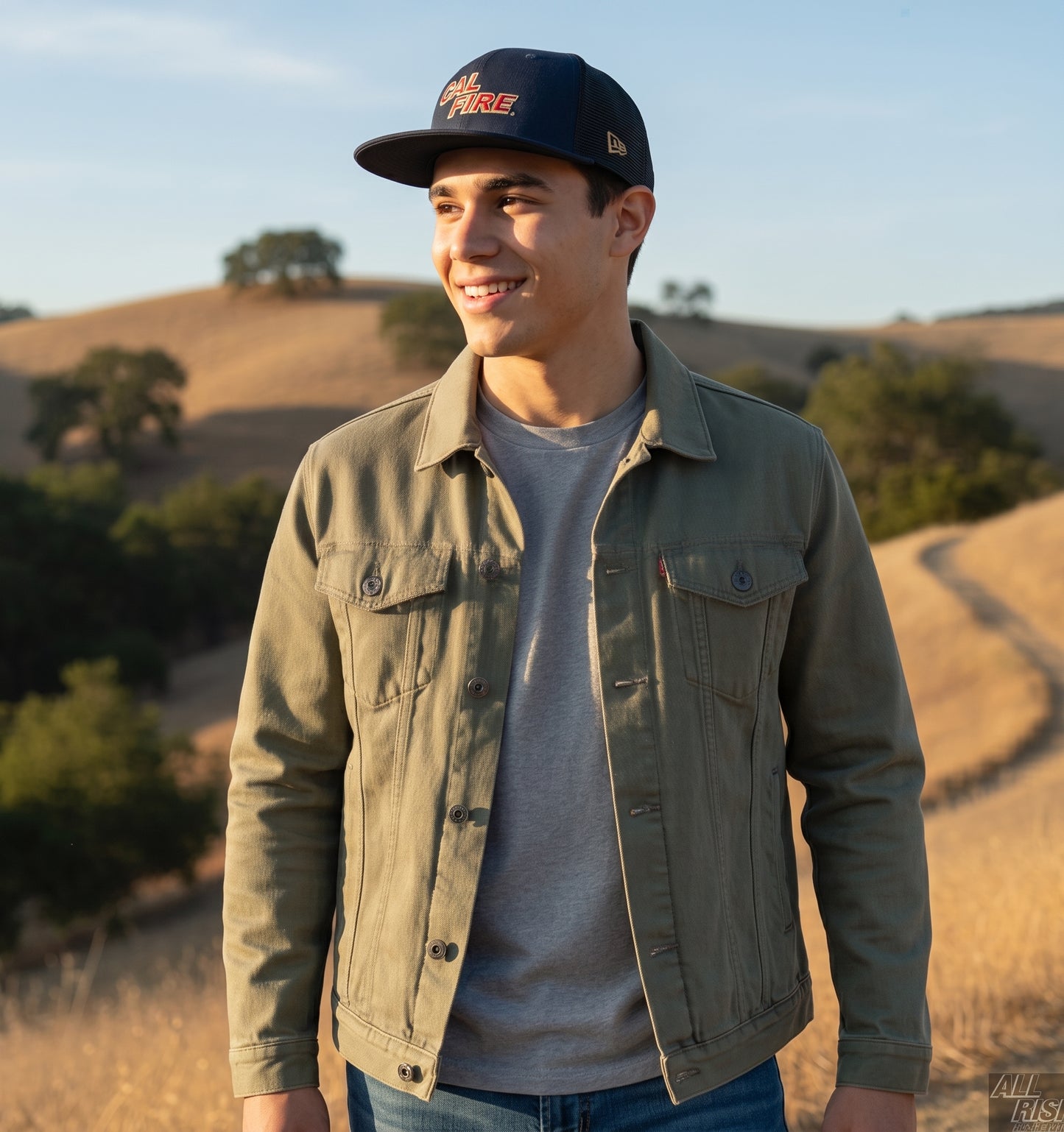 Lifestyle shot of a smiling young man in golden California foothills wearing the navy mesh-back CAL FIRE snapback with the red wordmark and an olive jacket.