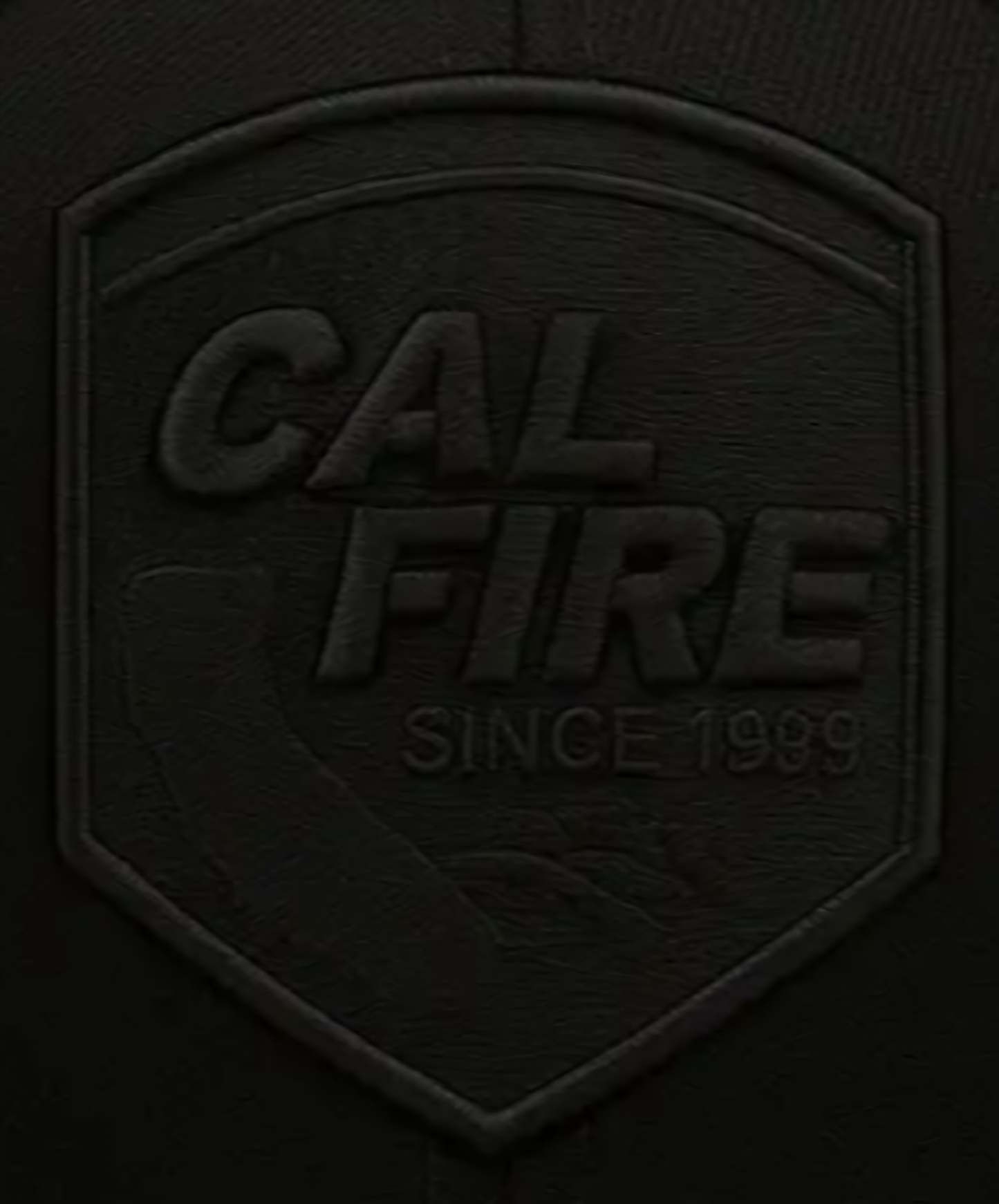 Macro close-up of the tonal black-on-black CAL FIRE shield crest on the all-black cap, with raised CAL FIRE SINCE 1999 stitching.