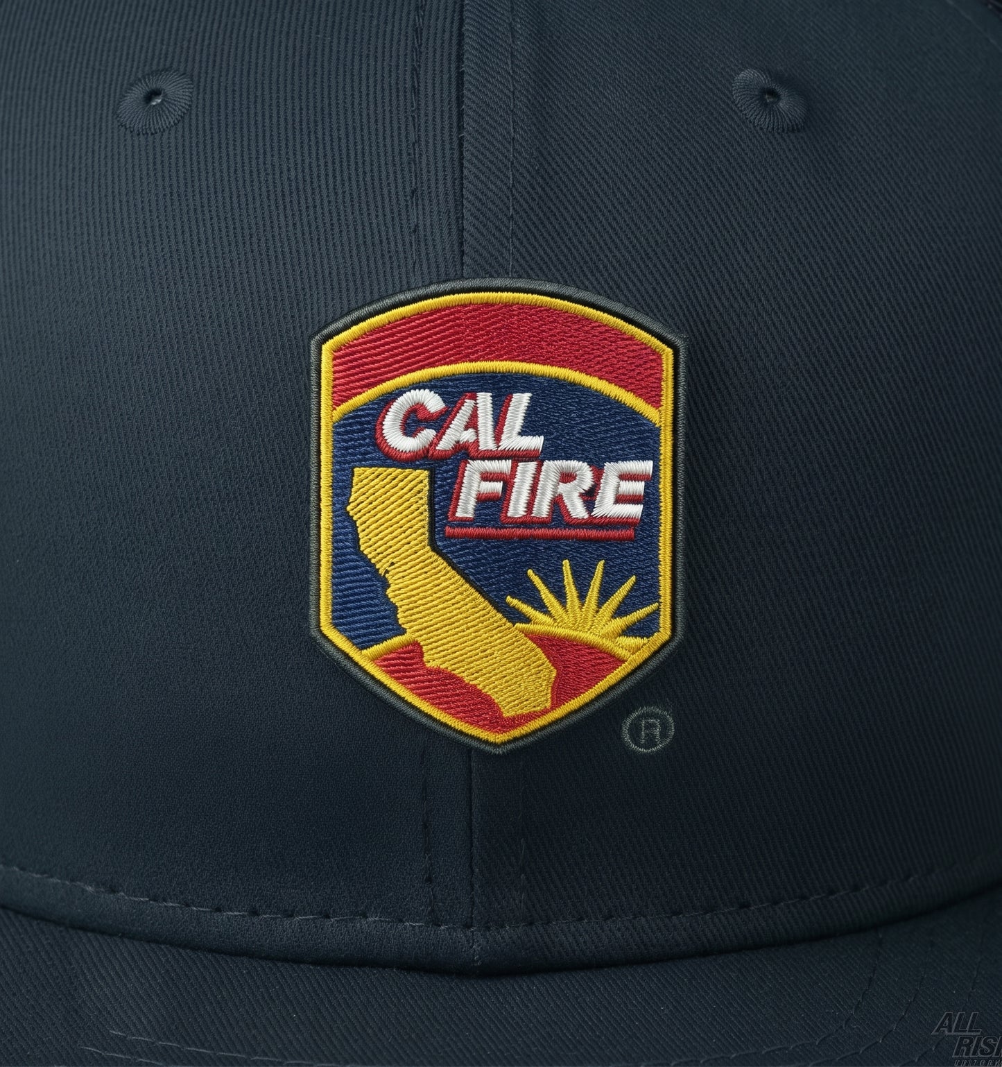 Close-up of the embroidered CAL FIRE arrowhead shield crest on the navy crown, showing the red and yellow shield, gold California outline and sunrise.
