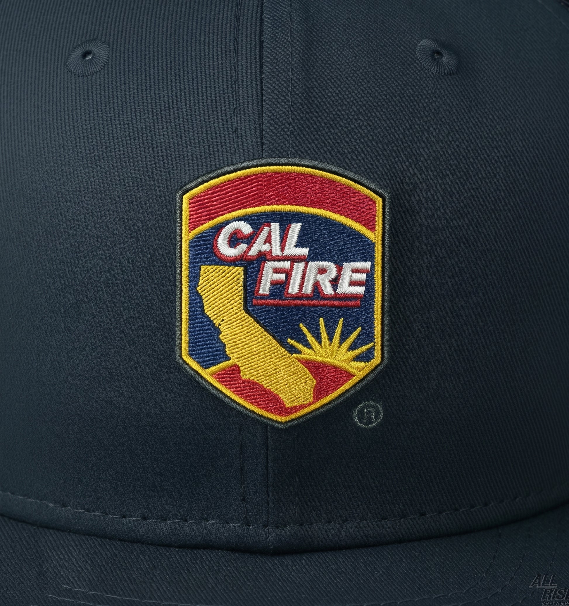 Close-up of the embroidered CAL FIRE arrowhead shield crest on the navy crown, showing the red and yellow shield, gold California outline and sunrise.